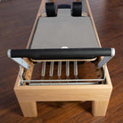 A wooden Pilates reformer machine with a gray reformer towel, metal foot bar, metal springs, and black shoulder pads, photographed on a dark wood floor.