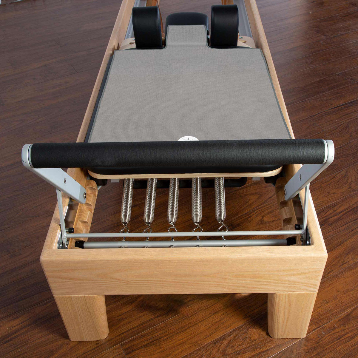 A wooden Pilates reformer machine with a gray reformer towel, metal foot bar, metal springs, and black shoulder pads, photographed on a dark wood floor.