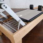 A close-up of a Pilates reformer machine showing the wooden frame,  metal foot rest, and black padded shoulder pads on a dark wood floor. Purple Reformer towel is on reformer carriage.