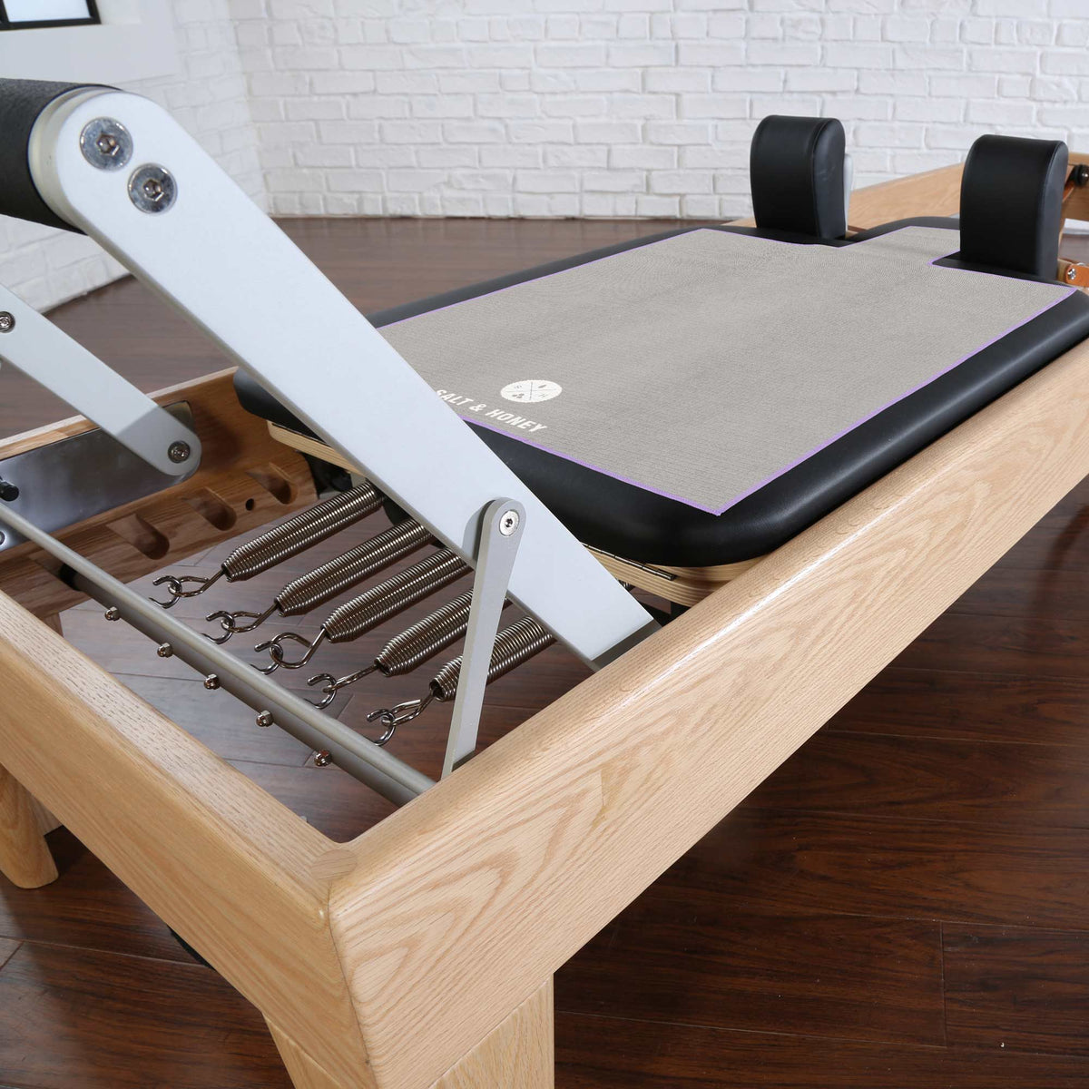 A close-up of a Pilates reformer machine showing the wooden frame,  metal foot rest, and black padded shoulder pads on a dark wood floor. Purple Reformer towel is on reformer carriage.